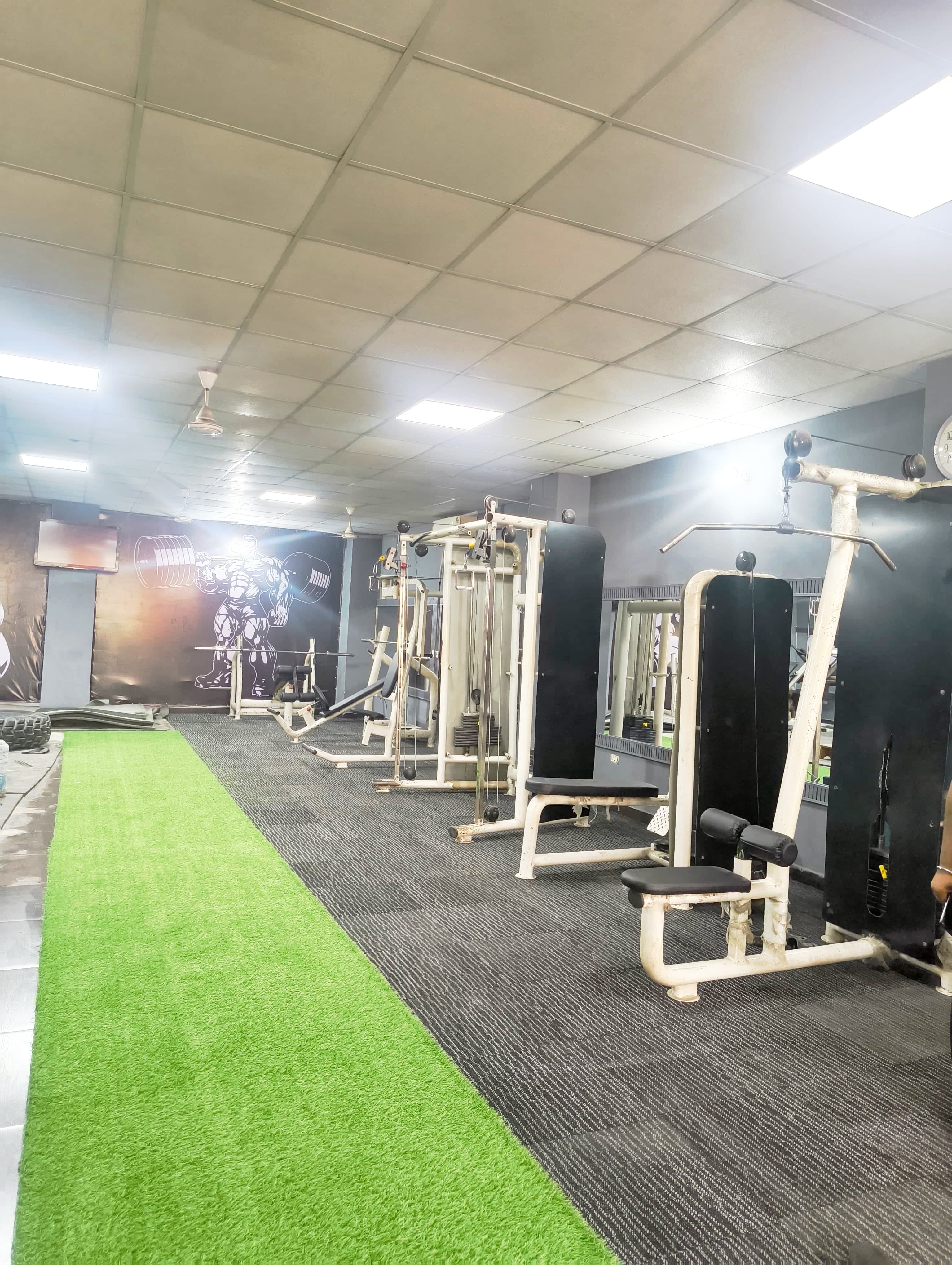 Gym interior featuring weight machines, a green turf strip, and a bodybuilder wall mural.